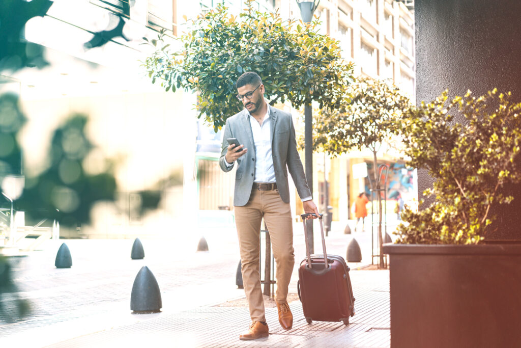 Image of a business traveler, walking with a suitcase towards his hotel, while looking at his phone