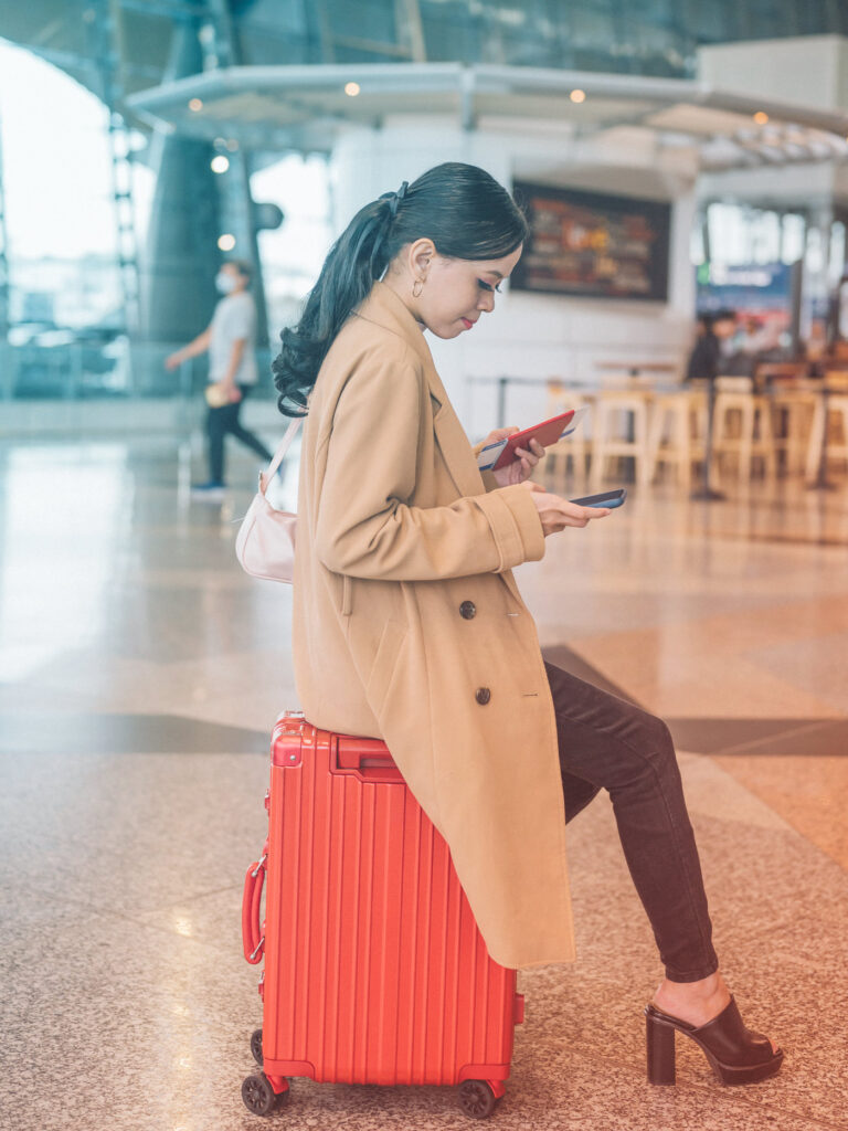 Image of a traveler, sitting on their suitcase in an airport, looking at their phone