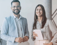 A man and wo,am dressed in business casual, standing next to each other smiling in an office environment.