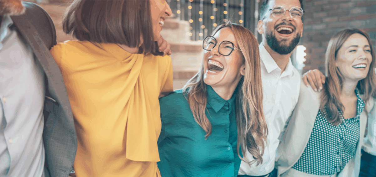 A group of diverse individuals stands together, all laughing joyfully. They are dressed in casual clothing and exhibit expressions of happiness and friendship.