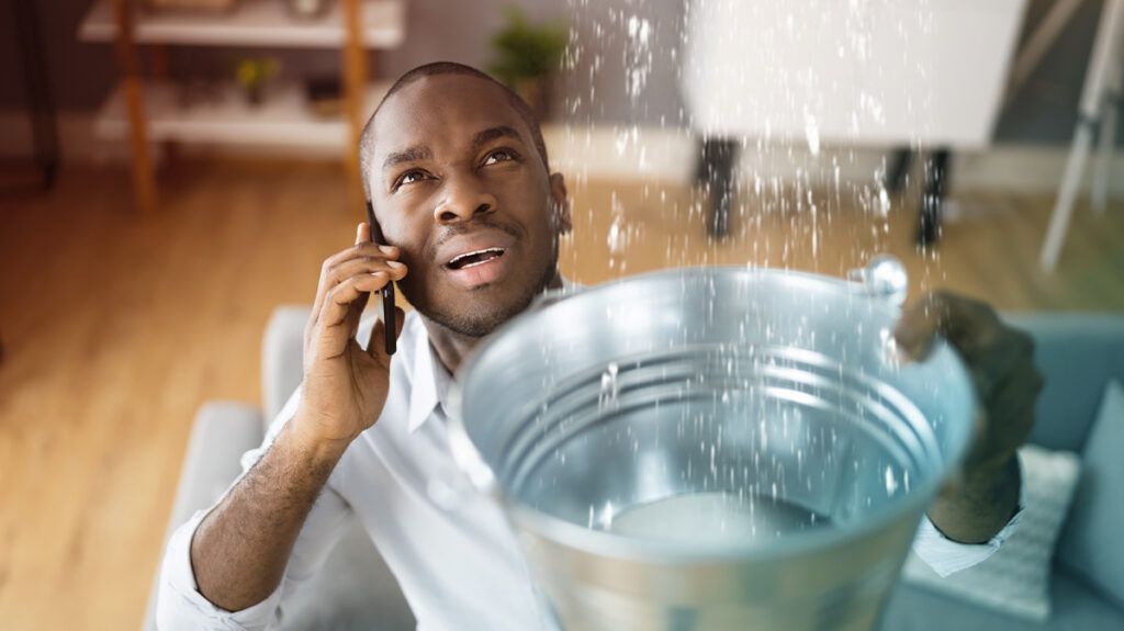 a man in distress on the phone, holding up a bucket catching water from a leaking ceiling dramatizing what is travel leakage